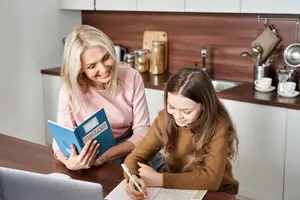 Woman and young girl sitting at a table in a kitchen, the woman is smiling and the girl is writing