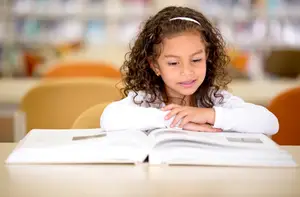 A girl with curly hair is sitting in a library reading a book.