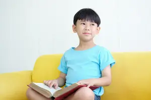 A smiling young boy with black hair sits on a yellow couch, holding a book in both hands.