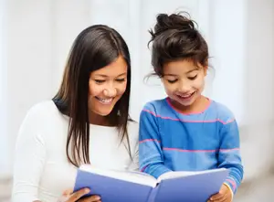 A woman and a girl are sitting together and reading a book. The woman is holding the book, and the girl is looking at it.