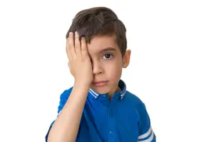 A young boy with short hair, wearing a blue polo shirt with white stripes, covering his face with his left hand, possibly feeling embarrassed.