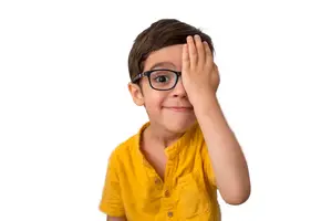 A young boy with glasses is covering his eye with his hand.