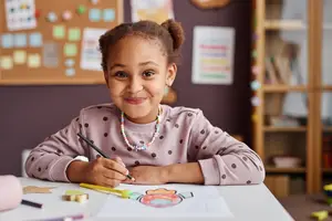 A smiling girl is sitting on a chair at a table, coloring a paper with a pencil and crayons in a classroom.