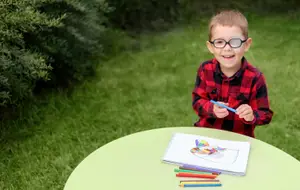 A little boy is smiling while sitting on a table and holding a blue pen in his hand. He is wearing a red and black checkered shirt and glasses. There is a green table with a notebook, a pen, and a pencil on it. There are also some colored pencils on the table. Behind him is a green lawn with some plants and bushes.