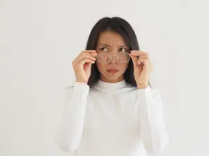 A woman wearing a white turtleneck shirt holds a pair of glasses in her hands, possibly adjusting them while looking at something.