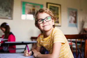 A young boy wearing glasses is smiling while holding a pen and doing an activity in the classroom