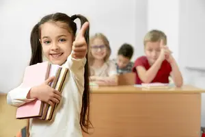 A girl holding books with a thumb up and a few other kids sitting behind her in a classroom.