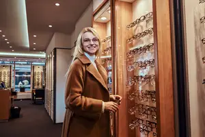 A woman in a brown coat is standing in front of a glass display case of eyeglasses in a store.