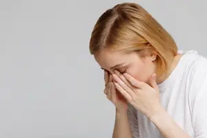 A woman with blonde hair wearing a white t-shirt appears to be sneezing while holding her nose and mouth with her hands.