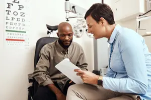A man with a bald head is sitting on a chair and having a conversation with a woman wearing a watch, and she is holding a paper and a pen. Behind them is a wall with an eye chart, and a chair with a machine is on the right side.