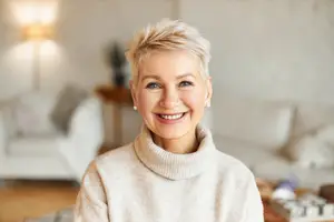 Portrait of a smiling mature woman with short hair and a turtleneck sweater, standing in a modern, bright living room.