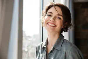 Portrait of a smiling woman with short hair wearing a denim shirt standing near a window