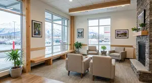A waiting room with wooden benches, chairs, and a fireplace. Potted plants are scattered around, and framed pictures adorn the walls. Outside the window, a snowy landscape with mountains and parked cars is visible.