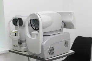 A white medical device with a circular lens and two monitors is on a desk in a white room.
