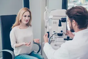 A woman sits in a chair while a doctor examines her eyes with a device.