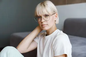 A young woman with short blonde hair wearing glasses and a white shirt sitting on a couch.