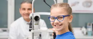 A girl wearing glasses is smiling while looking into an ophthalmoscope with a man in a white coat standing behind her in an eye clinic.