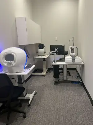 A medical examination room equipped with an advanced diagnostic machine, a computer monitor, and various medical devices, set against a white wall and gray carpet.