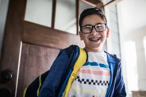 A young boy with glasses and a backpack is smiling and posing for a photo in front of a wooden door.
