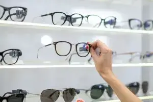 A person holding a pair of black framed glasses in a store with many other glasses on display