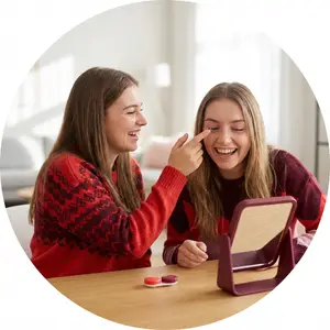 Two women sitting at a table with a small mirror, smiling and looking at each other with one putting a contact lens in the other's eye