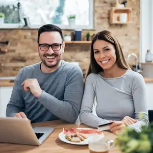 A couple is smiling while using a credit card on a laptop while sitting at a dining table with food and a cup of coffee in a kitchen.