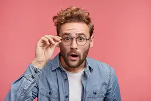 A man adjusts his glasses while standing against a pink background.
