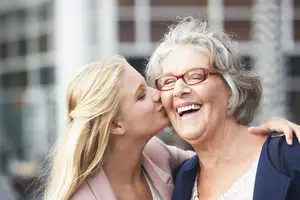 An elderly woman with gray hair and red glasses kissing a young woman with blonde hair