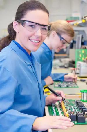 Two women wearing safety glasses are working on a circuit board in a factory