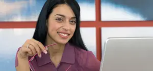 A smiling woman holding a pair of pink glasses in front of a laptop