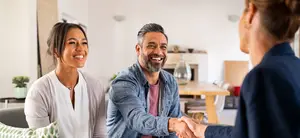 Three adults are in a living room with a woman in a suit shaking hands with a couple