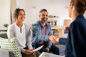Three adults are in a living room with a woman in a suit shaking hands with a couple