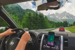 Driver driving on mountain road with mountains and lush greenery in the background, a red car in front, and a dashboard view with a cell phone, steering wheel, and speedometer.