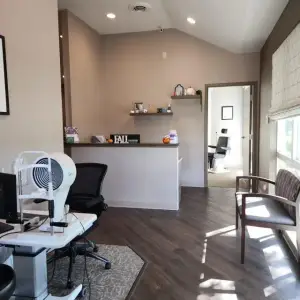 The interior of an optometrist's office featuring a white desk with an eye exam machine, a black swivel chair, a white reception desk, and a framed picture on the wall.