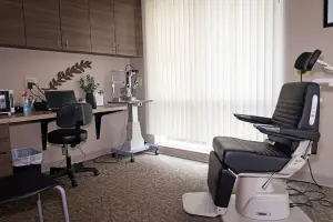 An empty optometrist examination room with a desk, a computer, a potted plant, and a chair.