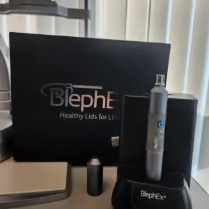 A BlephEx device is on a charger and beside a BlephEx box on a table with blinds in the background