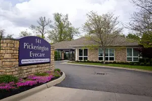 A stone sign with the words '141 Pickerington Eyecare' is on the left side of the building entrance, which has a brick wall, glass windows, and a door with a black frame. There are trees and bushes on the side of the building, and a street light is on the left side of the driveway. There are also plants and flowers on the side of the driveway, and the sky is cloudy.