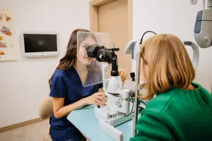 A woman with brown hair is checking the eyes of another woman with blonde hair using a medical machine in a room.