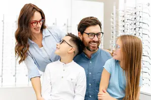 Four people are standing in a room, perhaps a store, with many eyeglasses hanging on the wall behind them. The woman is wearing glasses and smiling. The man is looking at something on the right. The boy and girl are looking up, perhaps at something. They are all wearing blue shirts. The boy is wearing a white shirt. The girl is wearing a blue shirt and has her hands on her waist.