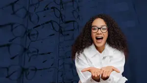 A smiling woman in glasses stands in front of a wall of glasses.