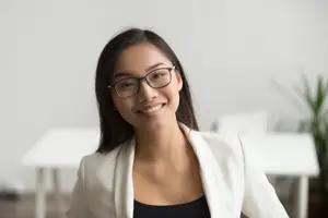 A smiling woman with glasses and a white blazer stands in an office with white walls and furniture.