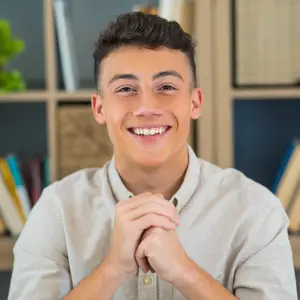A smiling young man is posing for a photo in a room with a wooden shelf behind him. He has his hands clasped together in front of his chest, and there is a potted plant on the left side of the shelf.