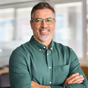 A man wearing glasses and a green polo shirt with his arms crossed and smiling while standing in an office.
