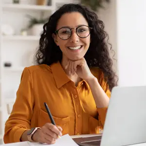 A smiling woman wearing glasses and a watch is sitting at a desk with a laptop and a pen in her hand.