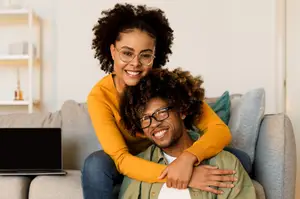 A smiling couple sitting on a couch with a laptop on the left side of the couch.