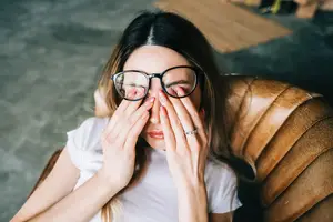 A woman wearing glasses and a white shirt is sitting on a brown leather couch and covering her eyes with her hands
