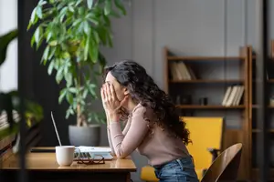 A woman is sitting at a desk in an office and is covering her face with her hands, maybe crying