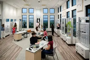 People inside an optical shop, some seated on chairs, and others standing near cabinets and looking at items on display.