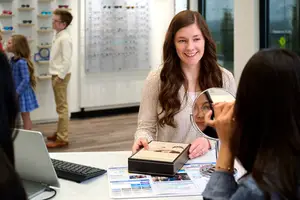 A woman is seen smiling while adjusting her glasses in front of a mirror, with a desk holding a laptop and a black box, and a woman on the right is adjusting her glasses, and a man and a girl are standing behind them in a room with shelves and a glass wall.