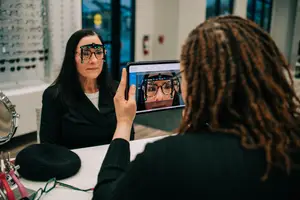 A woman is sitting in front of a table while a woman is holding a tablet in front of her face. They are both wearing glasses.